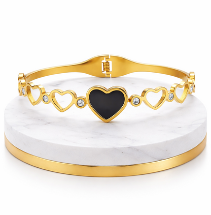 Gold bracelet with a black heart charm on a white marble stand